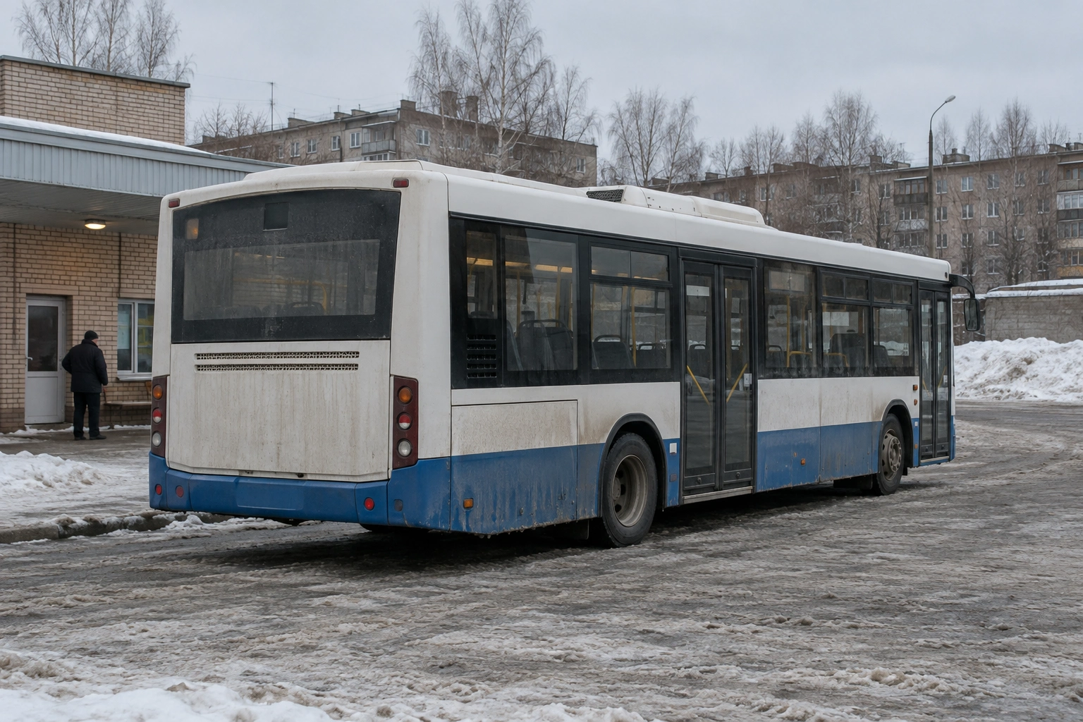 Городской автобус на заснеженной конечной остановке рядом с кирпичным служебным зданием.